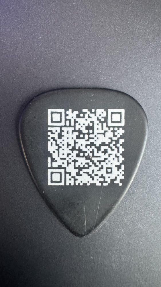 A black guitar pick with a white QR code printed on it, resting on a dark gray surface
